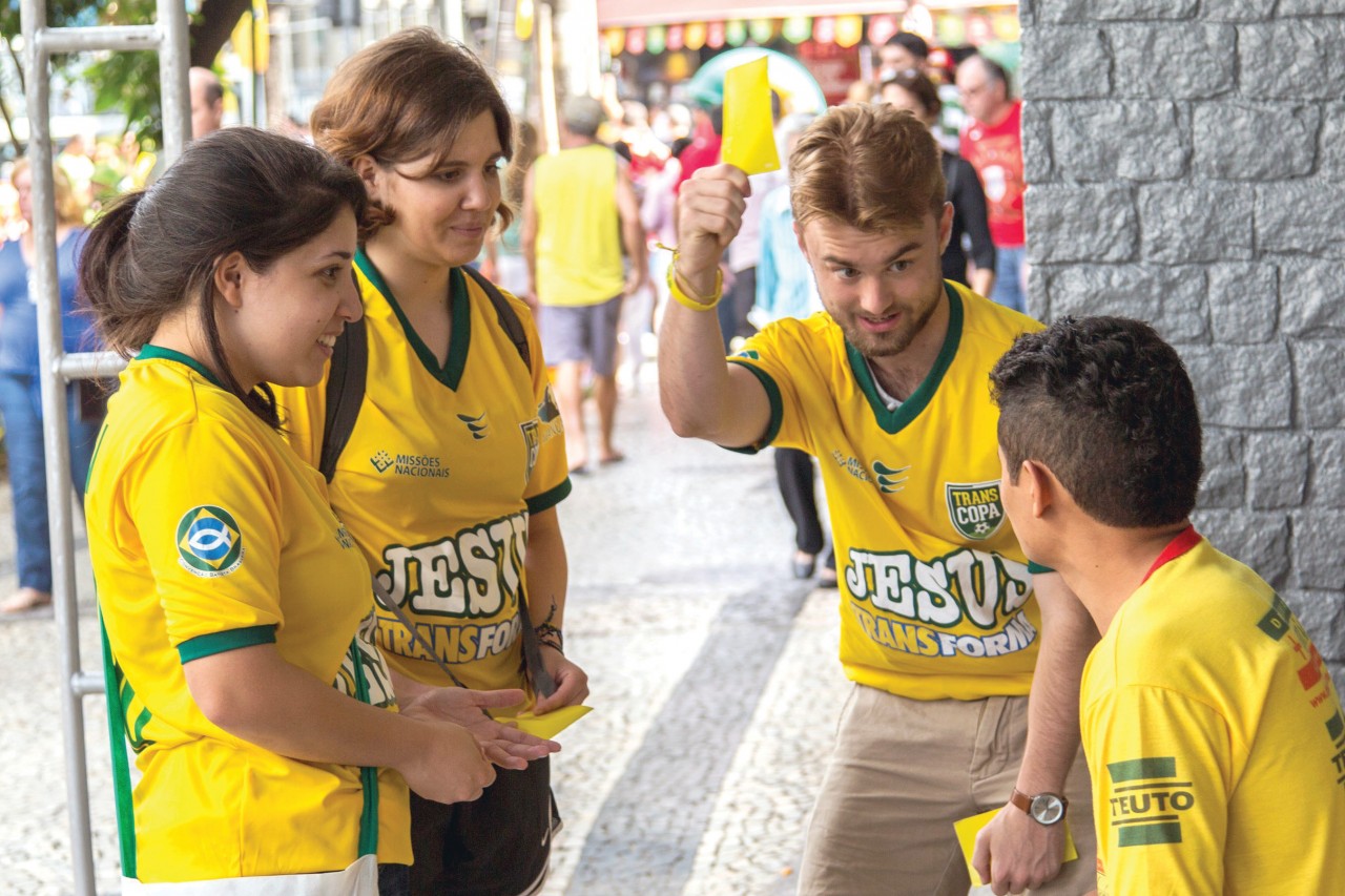 photo: Wilson  Hunter Student volunteer Grant Gilliam raises a yellow card—resembling what a referee uses to call a rule violation in a soccer match—to get the attention of a soccer fan near Rio de Janeiro’s Maracana Stadium. Gilliam and his teammates, from left, Bianca Brandao and Stephanie Silva, a student at Oklahoma Baptist University, shared the Gospel before a World Cup match.
