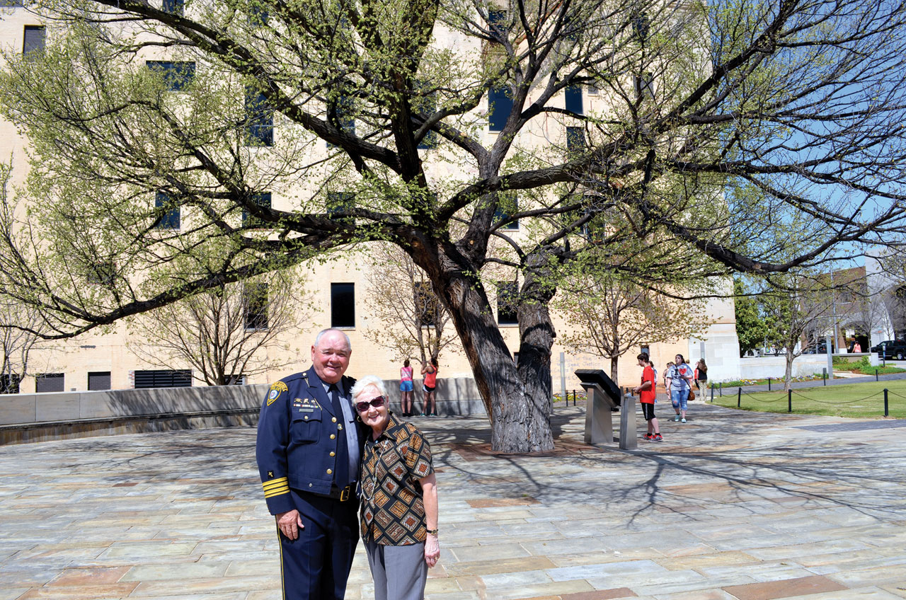 Jack and Phyllis Poe pose in front of The Survivor Tree, an American Elm that stood in the parking lot of the Journal Record Building, now the Oklahoma City National Memorial, shown in the background. (Photo: Bob Nigh)